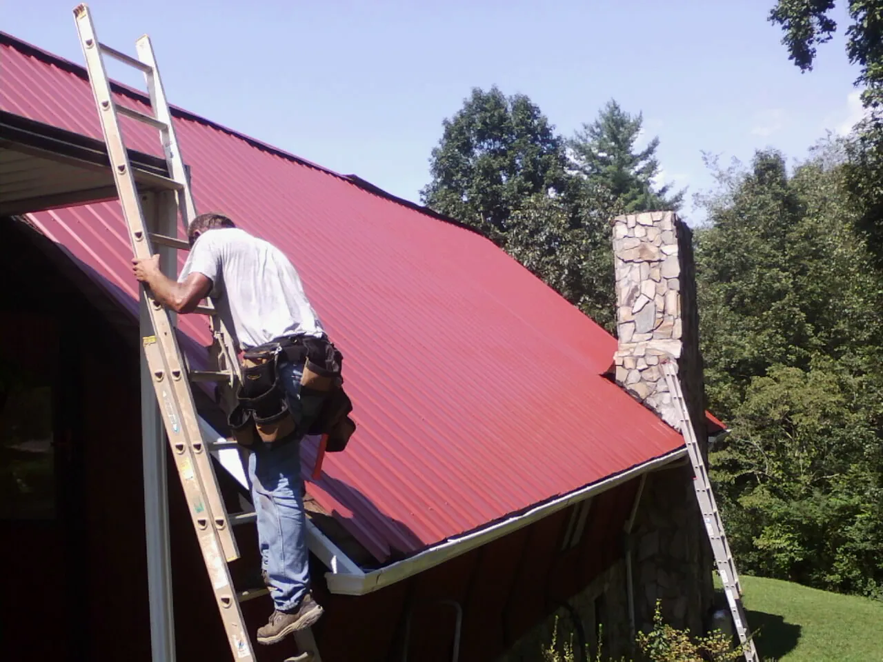 Roofer on a ladder beside a red metal roof and stone chimney — Soffit & Fascia Repair by Roofing Brothers in Hustonville